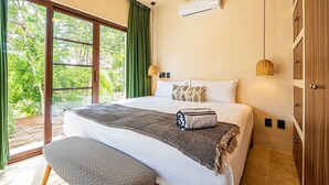 Un dormitorio con una cama grande, un banco, aire acondicionado de pared y vistas a zonas verdes a través de la ventana.