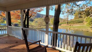 A covered porch with wooden flooring and white railings, overlooking a body of water and autumn foliage. - Family-sized Cozy Cottage - mountains views, fishing, & hot tub, in Suches, GA (Suches)