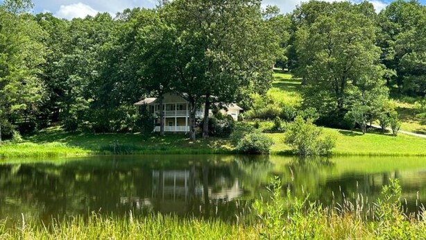 A house with a pond in front, surrounded by lush greenery and trees. - Family-sized Cozy Cottage - mountains views, fishing, & hot tub, in Suches, GA (Suches)