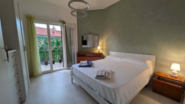 Un dormitorio con cama, mesita de noche, espejo, ventana con cortinas y vista a un edificio de techos rojos.