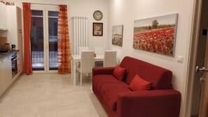 Un salón con un sofá rojo, una mesa de comedor con sillas, una zona de cocina y un amplio ventanal con persianas.
