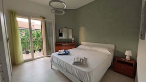 Un dormitorio con cama, mesita de noche, cómoda, espejo y puerta corrediza de vidrio que da a un balcón con plantas en macetas.