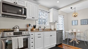 Cocina moderna con armarios blancos, electrodomésticos de acero inoxidable y una zona de comedor con sillas transparentes y mesa de madera.