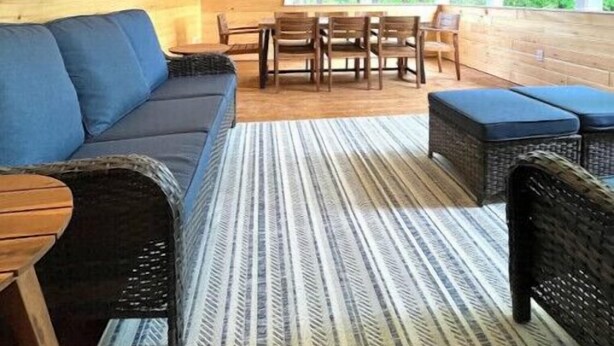 A cozy cabin interior with a wooden ceiling, a blue sofa, wicker furniture, and a large window offering a view of greenery. - 4BD 4.5BA Pet-Friendly Riverfront Home (Saco)