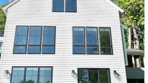 A two-story house with large windows and a covered patio area with Adirondack chairs. - 4BD 4.5BA Pet-Friendly Riverfront Home (Saco)