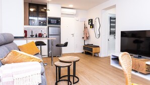 Una sala de estar moderna con un sofá, dos mesas redondas, un televisor y una zona de cocina con refrigerador y horno.