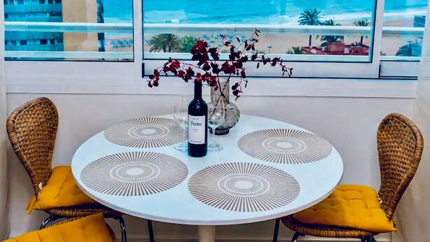 Una mesa redonda con una botella de vino, dos sillas de mimbre con cojines amarillos y vista al océano a través de amplios ventanales.