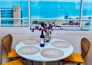 Una mesa redonda con una botella de vino, dos sillas de mimbre con cojines amarillos y vista al océano a través de amplios ventanales.