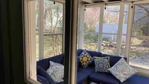 A room with a dark blue sofa set against a white wall, a window with a view of a yard, and a small table with a tray of orange flowers.