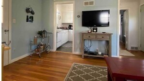 A living room with a wooden floor, a TV on a stand, and a kitchen in the background.