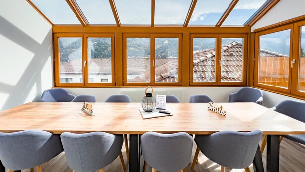 Une salle de conférence dotée d’une grande table en bois et de six chaises grises. Elle est agrémentée d’une verrière avec de nombreuses fenêtres offrant une vue sur un quartier résidentiel aux toits recouverts de tuiles.