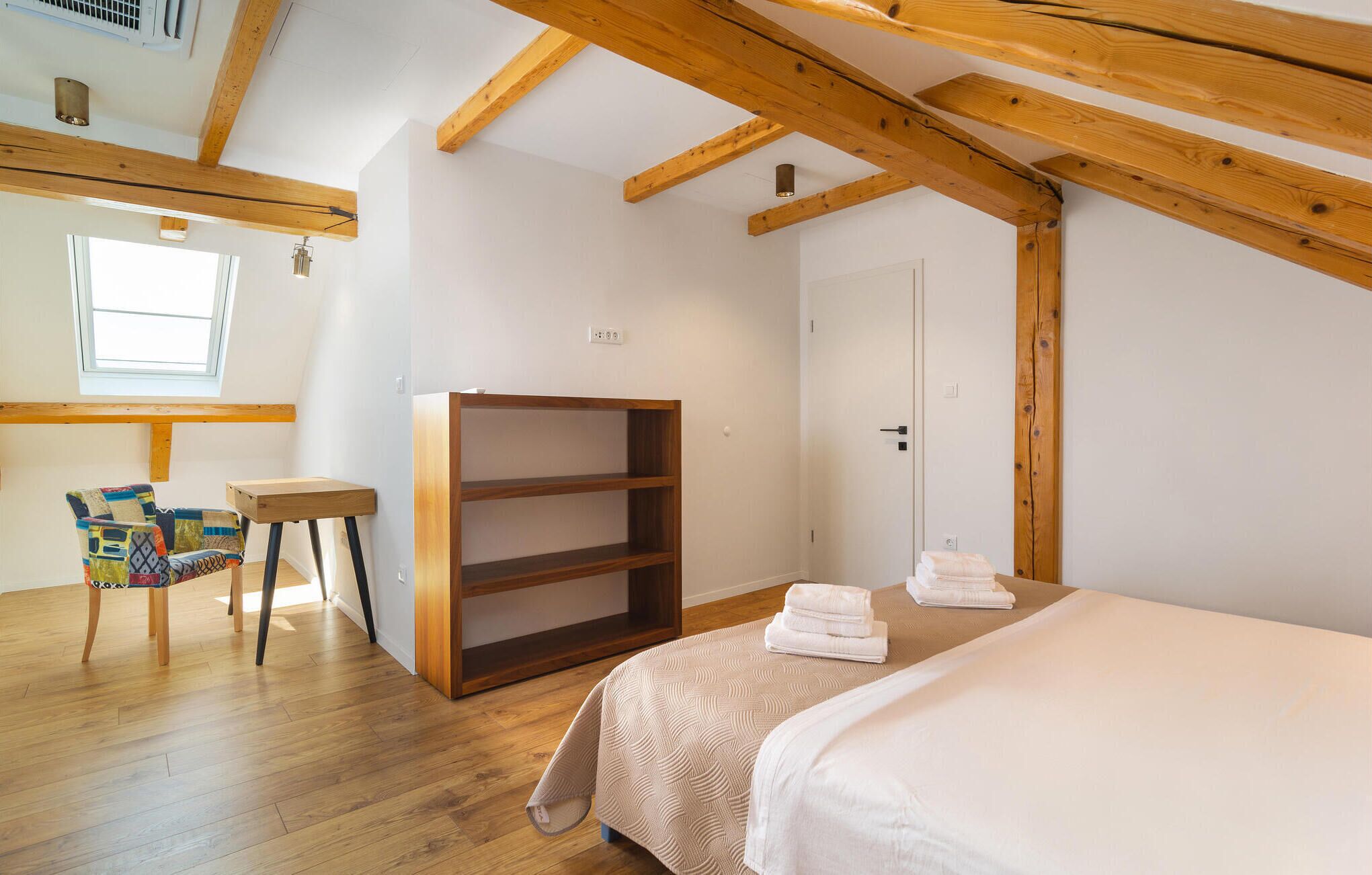 A bedroom with a bed, a wooden wardrobe, a small table, a chair, and a window.