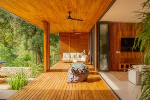 Um deck de madeira com sofá, poltrona de saco de feijão, ventilador de teto e vista para o verde.