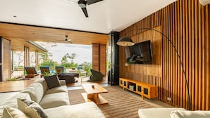 A modern living room with a large sofa, a coffee table, and a TV mounted on a wooden wall. There is a ceiling fan and a view of the outdoors through large windows. - The L Design Villa (Santa Teresa de Cobano)