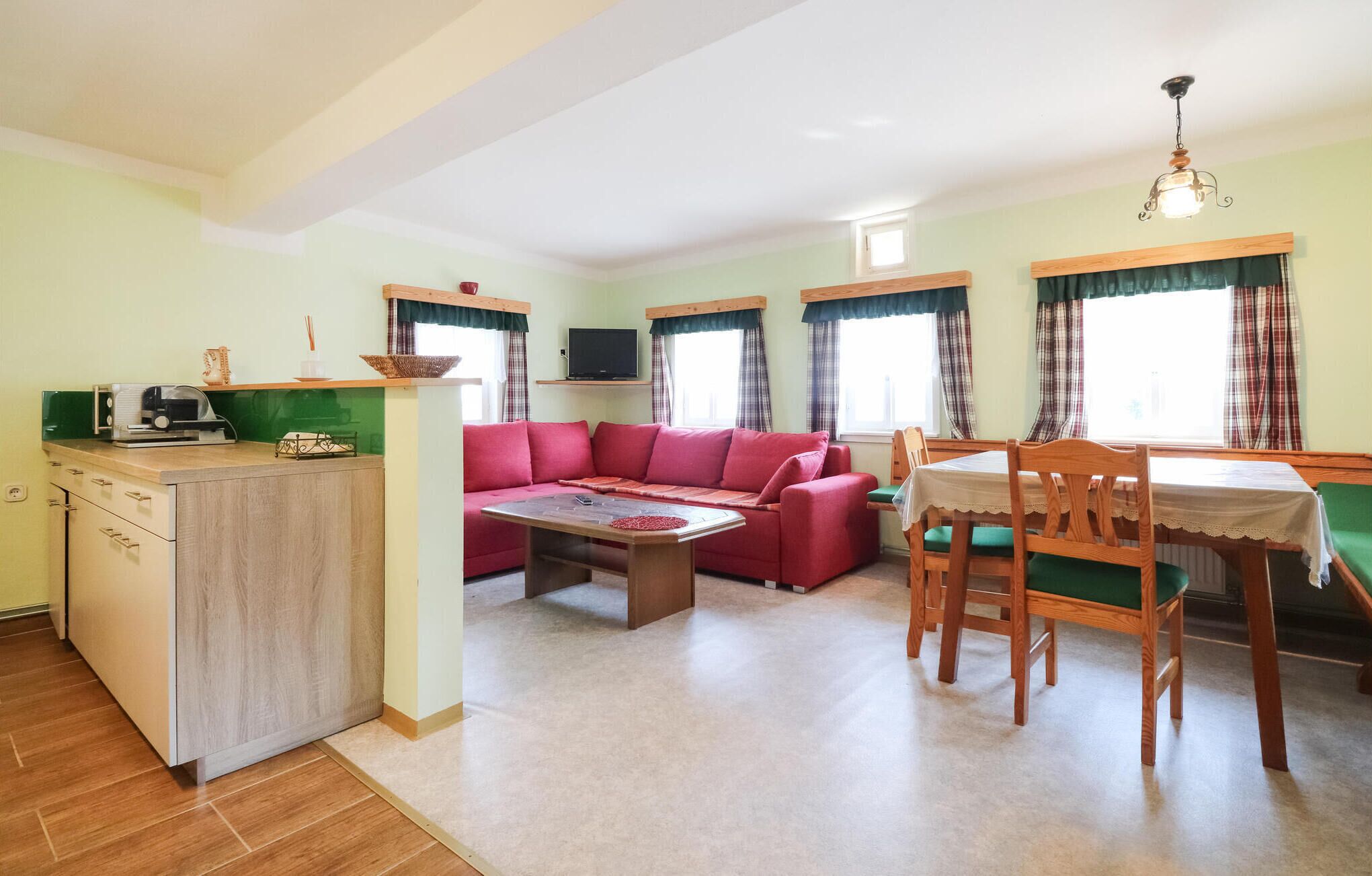 A cozy living room with a red sofa, a wooden dining table with chairs, and a kitchen area with a countertop and cabinets. - Ferienhaus mit Landblick (Oberhaag)