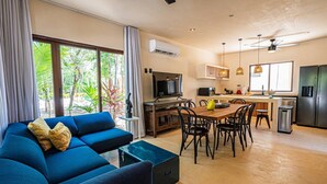 Un salon moderne avec un canapé bleu, un coin repas avec une table et des chaises en bois, une cuisine équipée d’un réfrigérateur et d’une cuisinière, et de grandes fenêtres avec des rideaux.