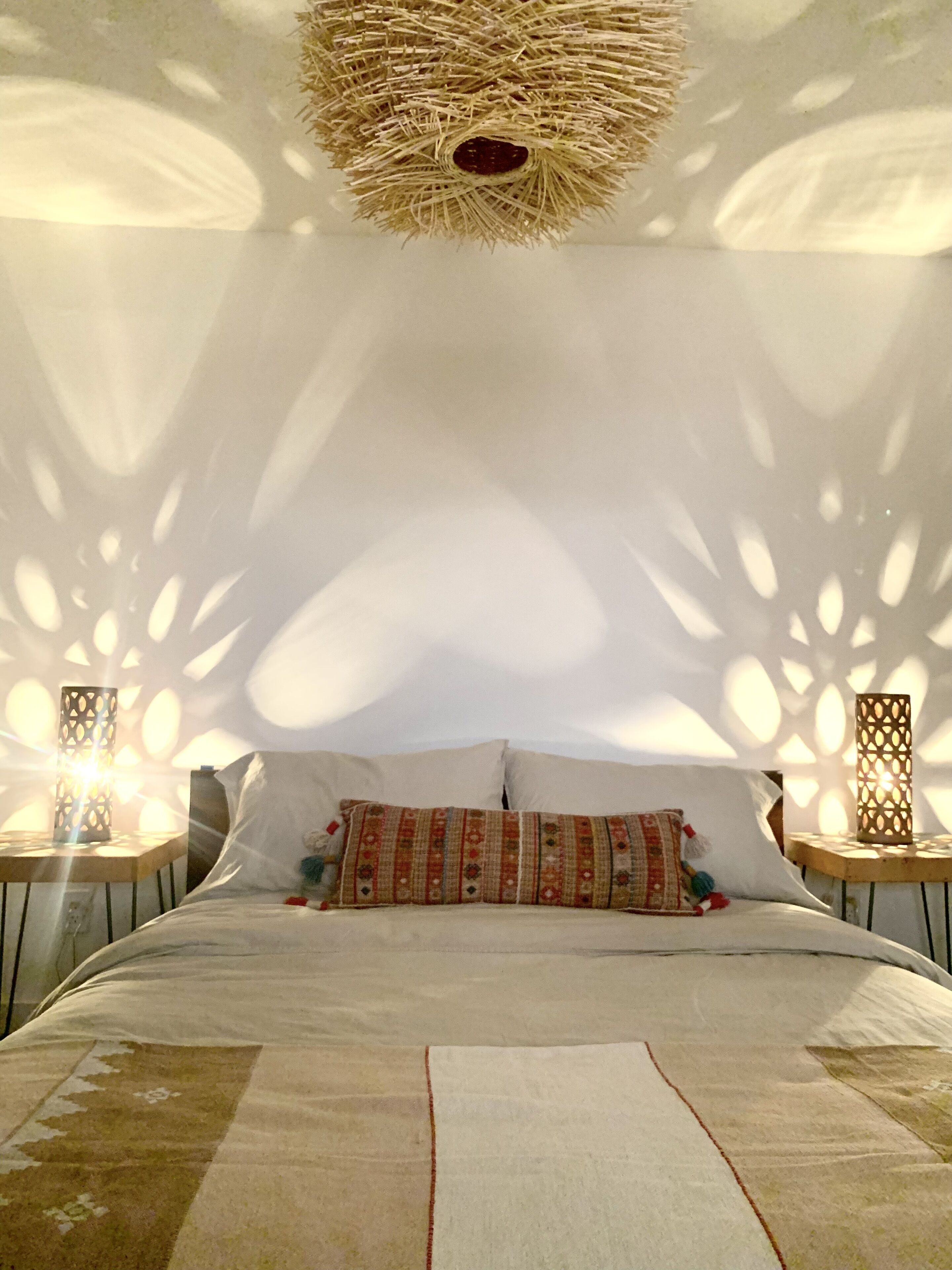 Uma cama com almofadas estampadas, duas mesas de cabeceira com abajures e um lustre decorativo.