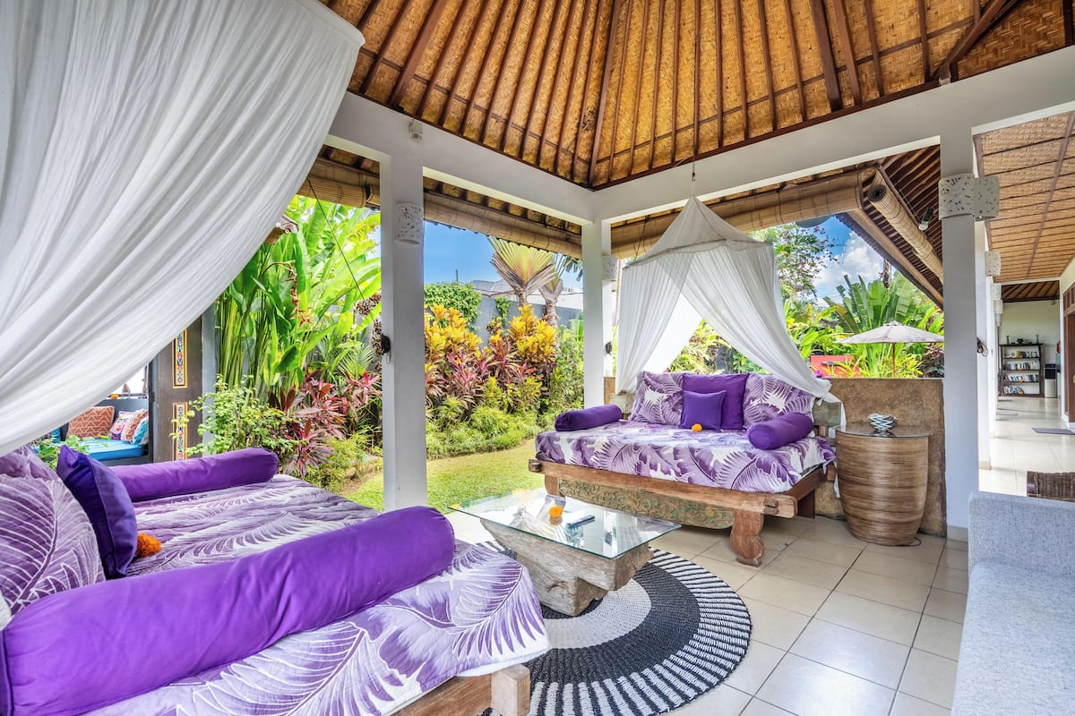 A covered outdoor seating area with a thatched roof, furnished with purple and white patterned sofas and a glass coffee table.