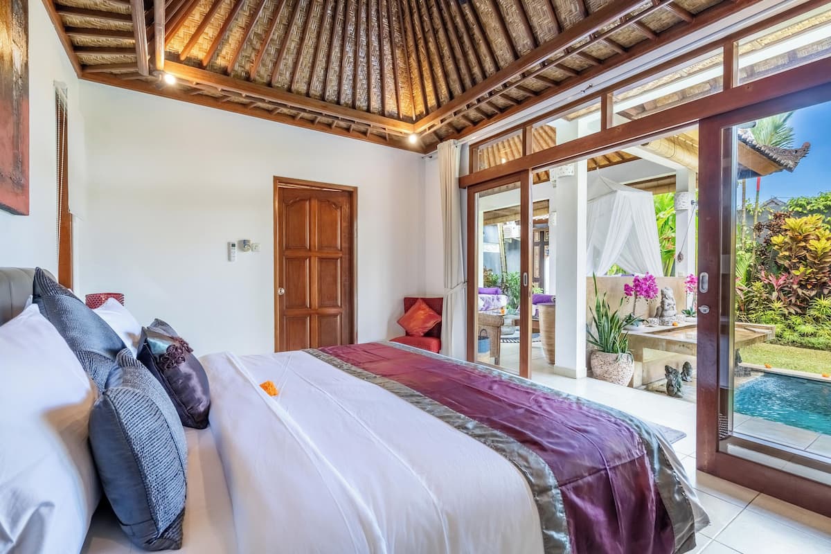 A bedroom with a large bed, a wooden ceiling, a sliding glass door leading to a pool area.