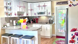 A modern kitchen with white cabinetry, a central island with bar stools, a microwave, and a refrigerator with floral decorations. - Boutique Guest House – Private Patio Oasis – Near Beach – Family-Friendly (North Miami)