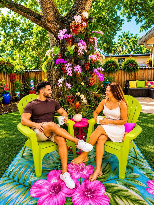 Two people seated on green chairs with a tropical-themed tablecloth, surrounded by potted plants and flowers.