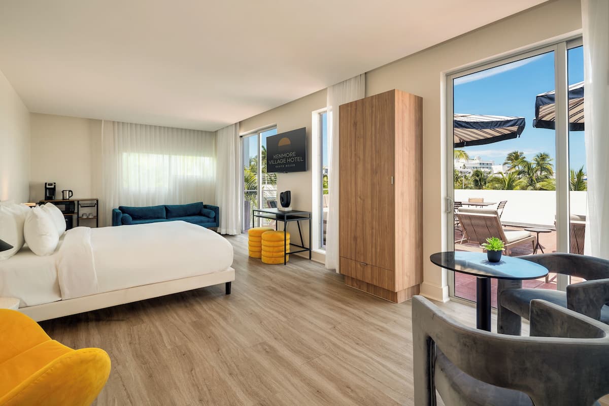 A hotel room with a bed, a desk, a chair, a small table with a plant, and a view of the outside.