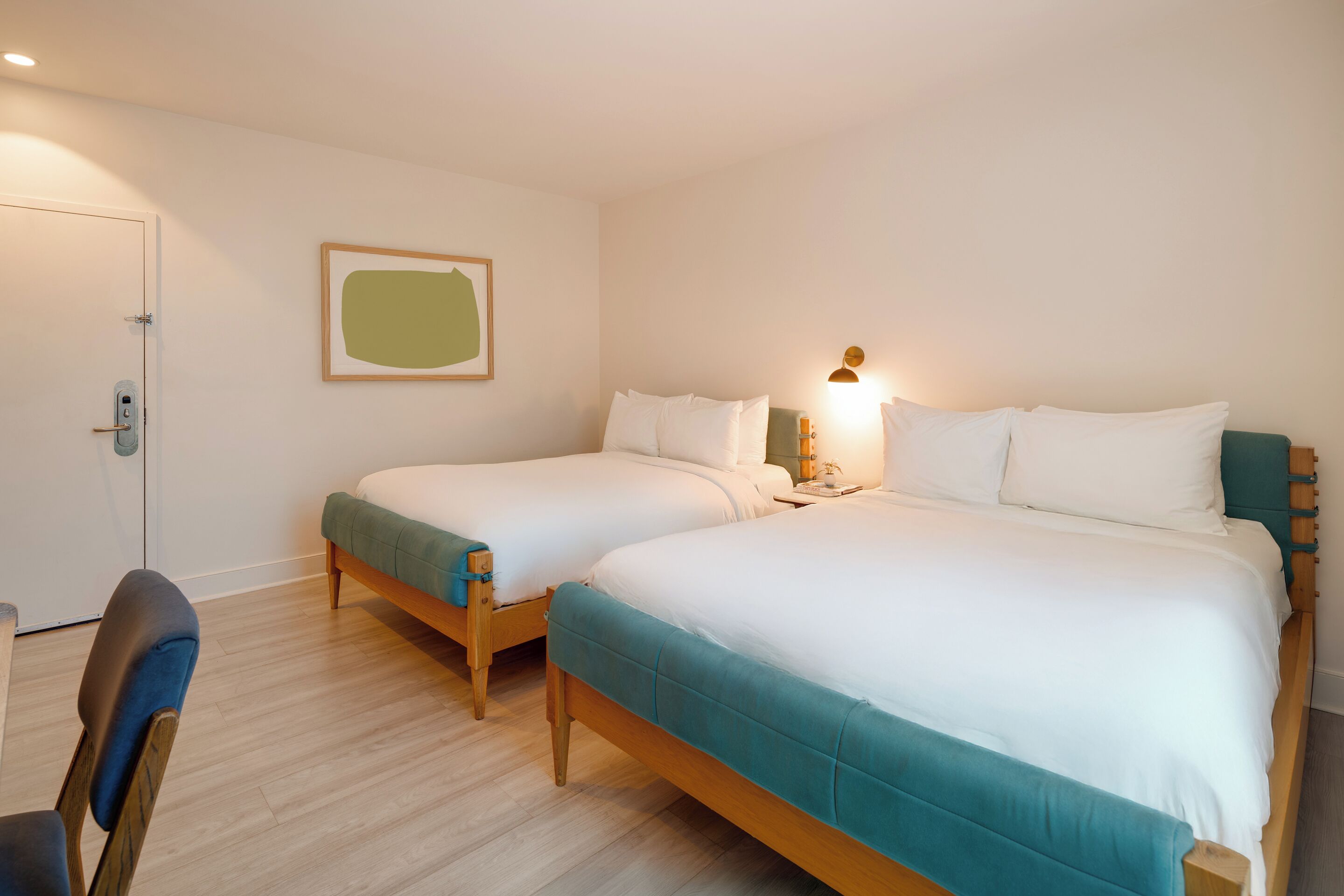 Une chambre d’hôtel avec deux lits simples, une chaise en bois, une porte avec une poignée et un luminaire fixé au mur.