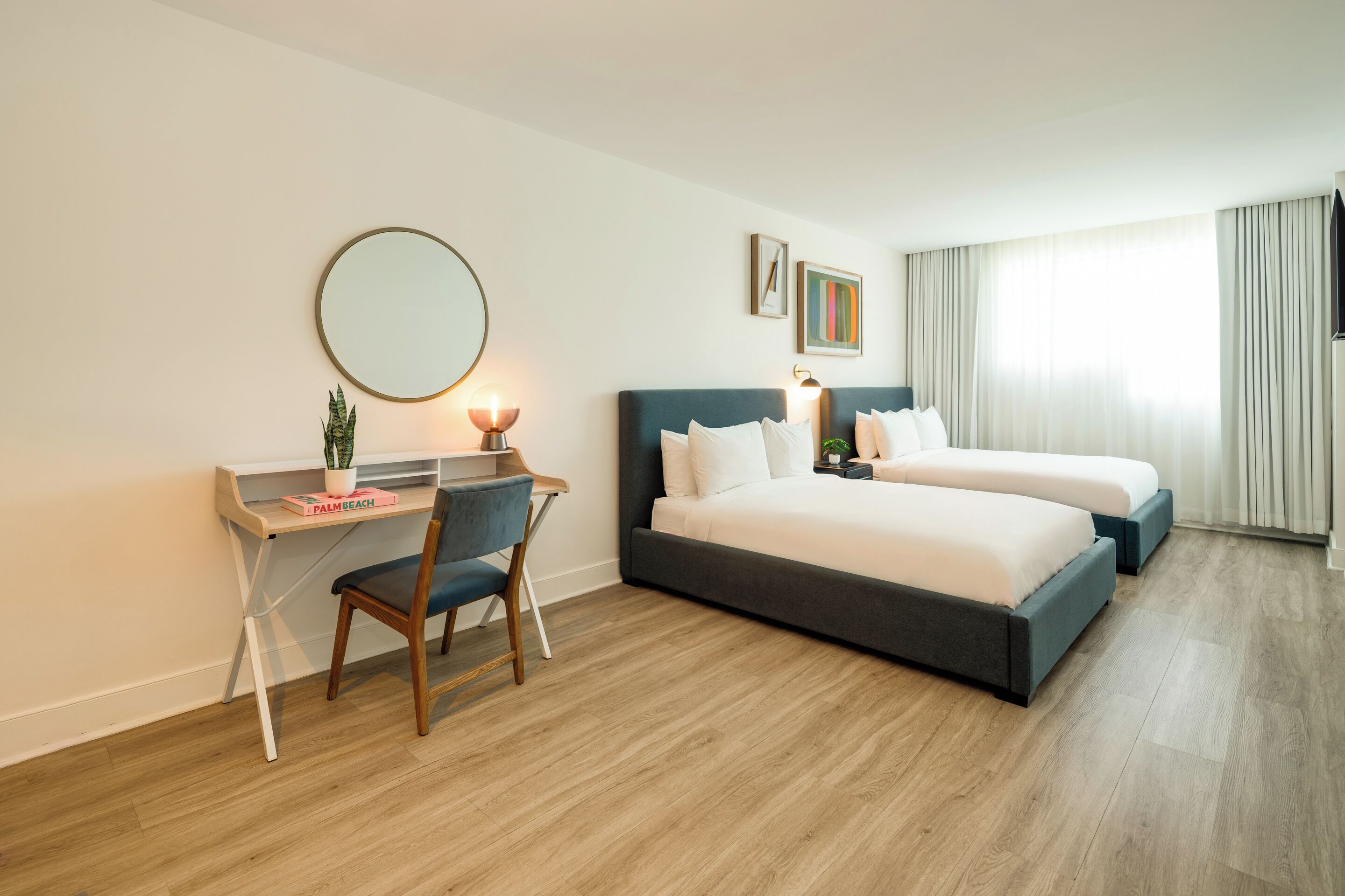 A hotel room with a desk, two beds, a round mirror, and a large window with curtains.