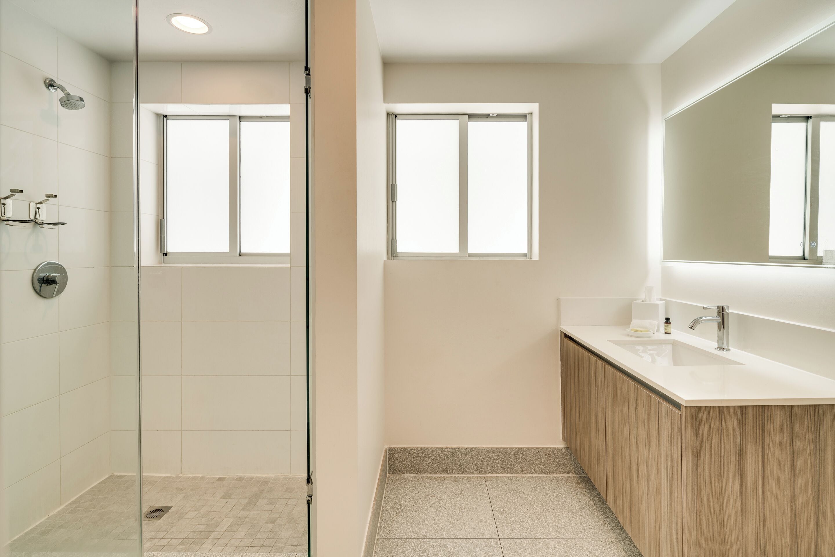 A modern bathroom with a glass shower enclosure, two windows, a wooden vanity, and a large mirror.