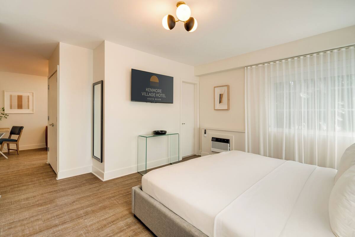 A hotel room with a large bed, a glass side table, a framed sign on the wall, and a window with curtains.
