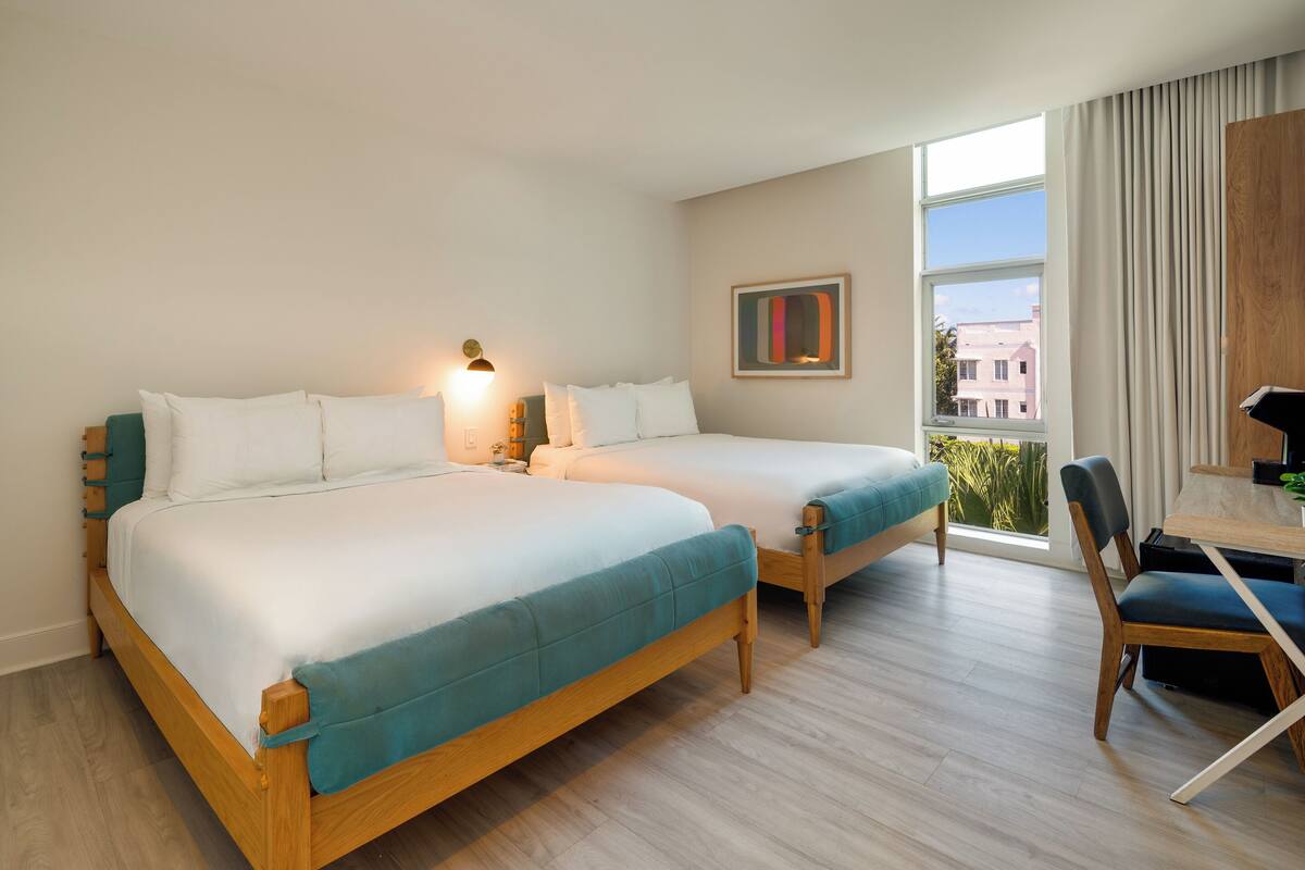 A hotel room with two beds, a desk, and a chair. There is a window with curtains and a view of buildings outside.