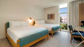 A hotel room with two beds, a desk, and a chair. There is a window with curtains and a view of buildings outside.
