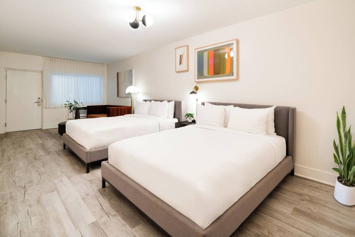 A hotel room with two beds, a large window with curtains, a potted plant, and a painting on the wall.