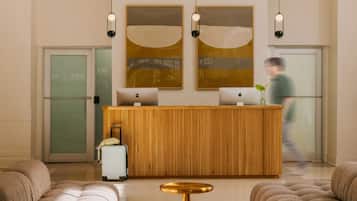A reception area with a wooden desk, two Apple computers, a plant, and a suitcase.