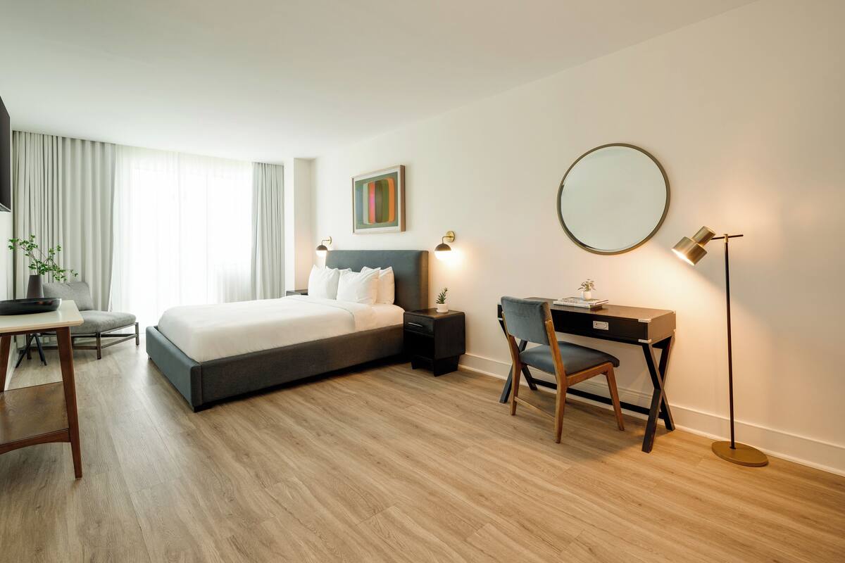 A modern hotel room with a large bed, a desk, a chair, a desk lamp, a round mirror, and a potted plant.