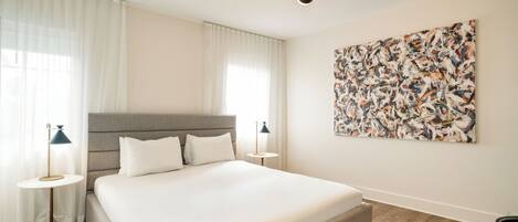 A modern hotel room with a large bed, two bedside tables with lamps, a large abstract painting on the wall, and a wooden floor.