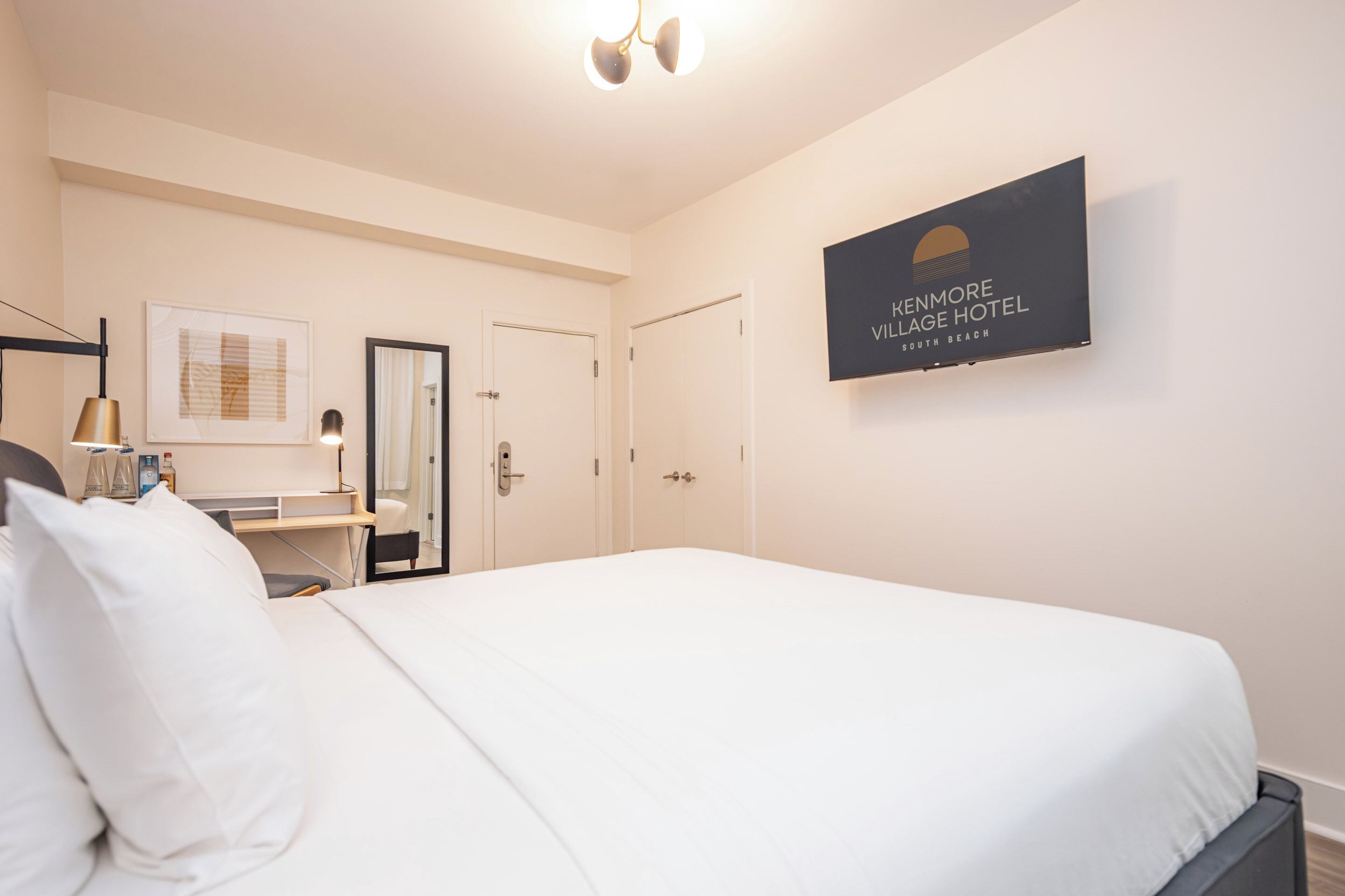 A hotel room with a large bed, a desk with a lamp, a framed picture on the wall, and a sign for Kenmore Village Hotel.