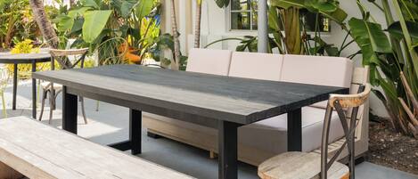 Una mesa de madera con bancos, un sofá acolchado y una silla en un espacio exterior con plantas tropicales.