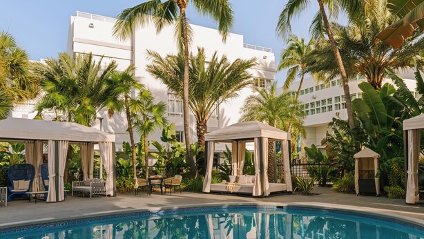 A pool area with a clear blue pool, surrounded by palm trees and covered seating areas. - Kenmore Village Hotel, South Beach (Miami Beach)