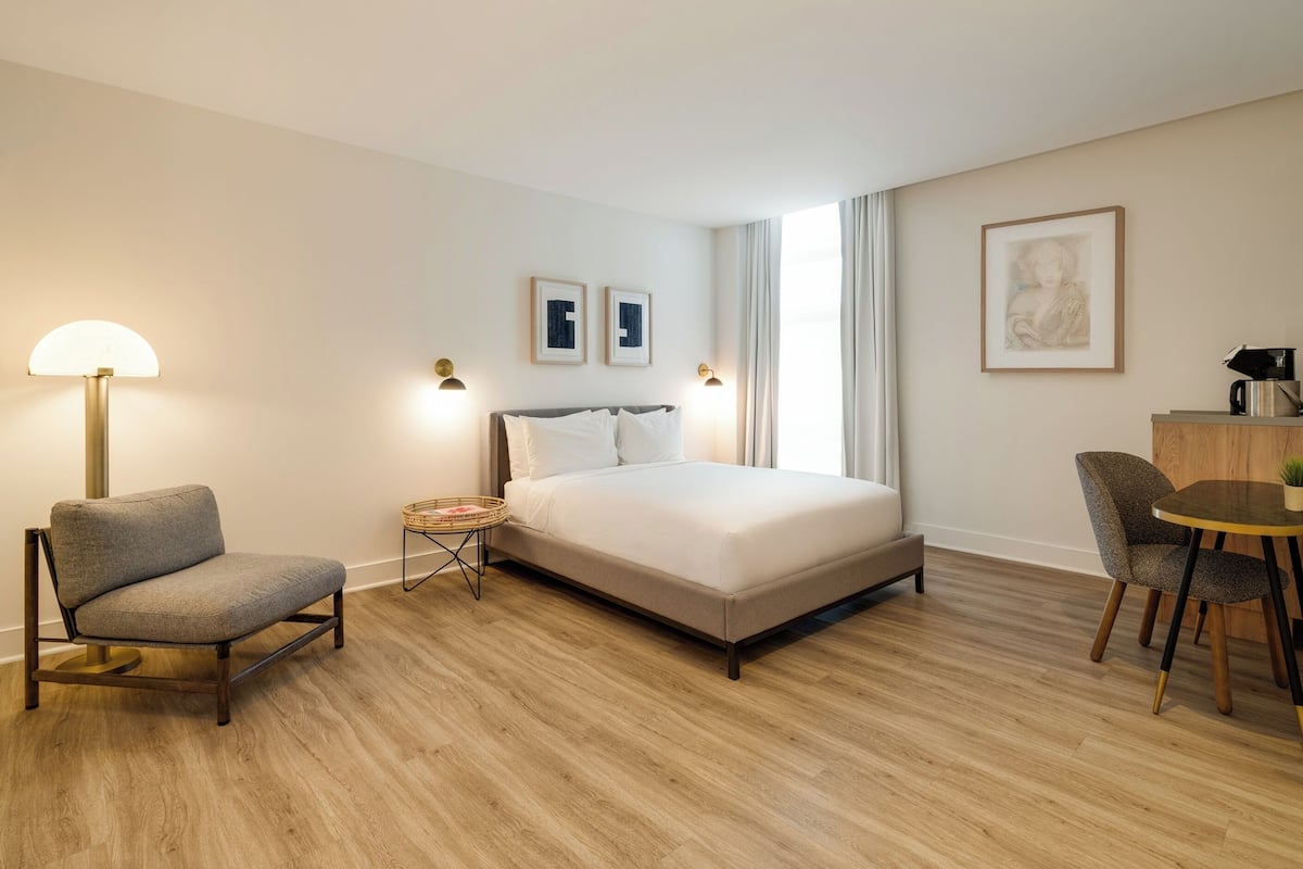 A hotel room with a bed, a chair, a small table, a lamp, and framed pictures on the wall.