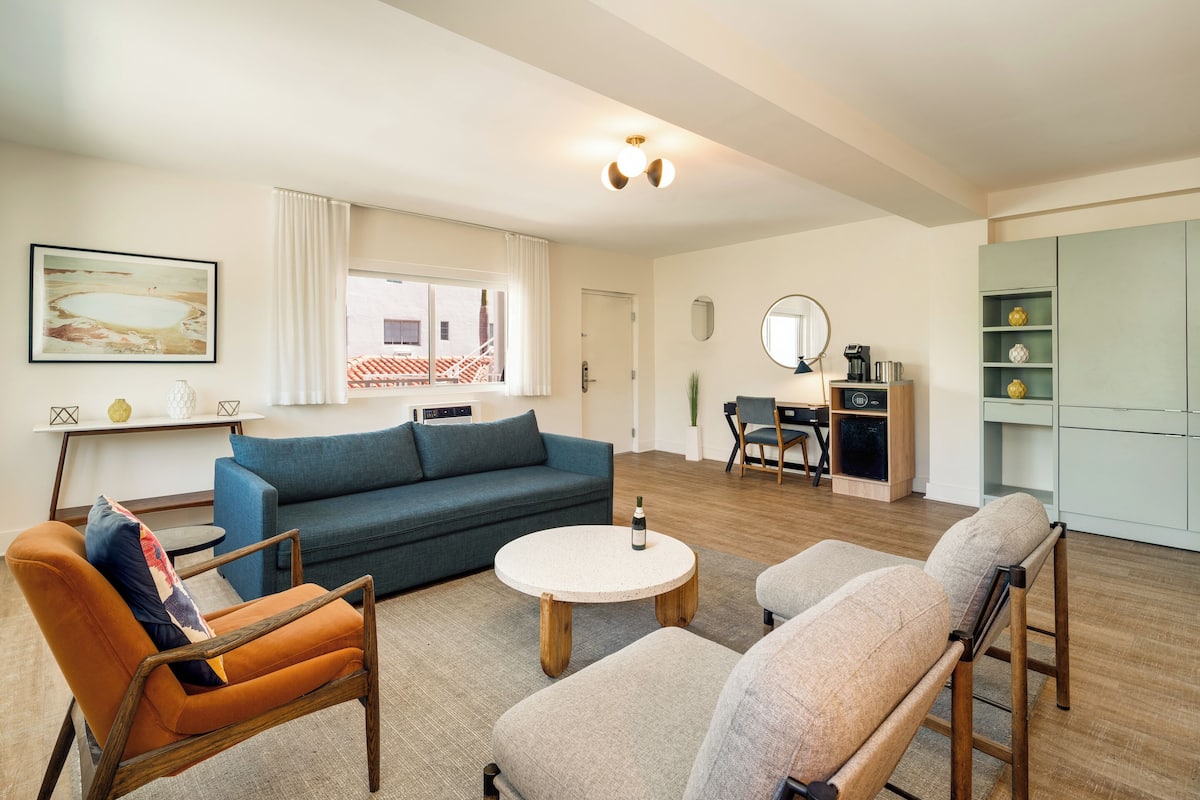A living room with a blue sofa, orange armchair, and beige armchair, a round coffee table, and a dining area with a table and chairs.