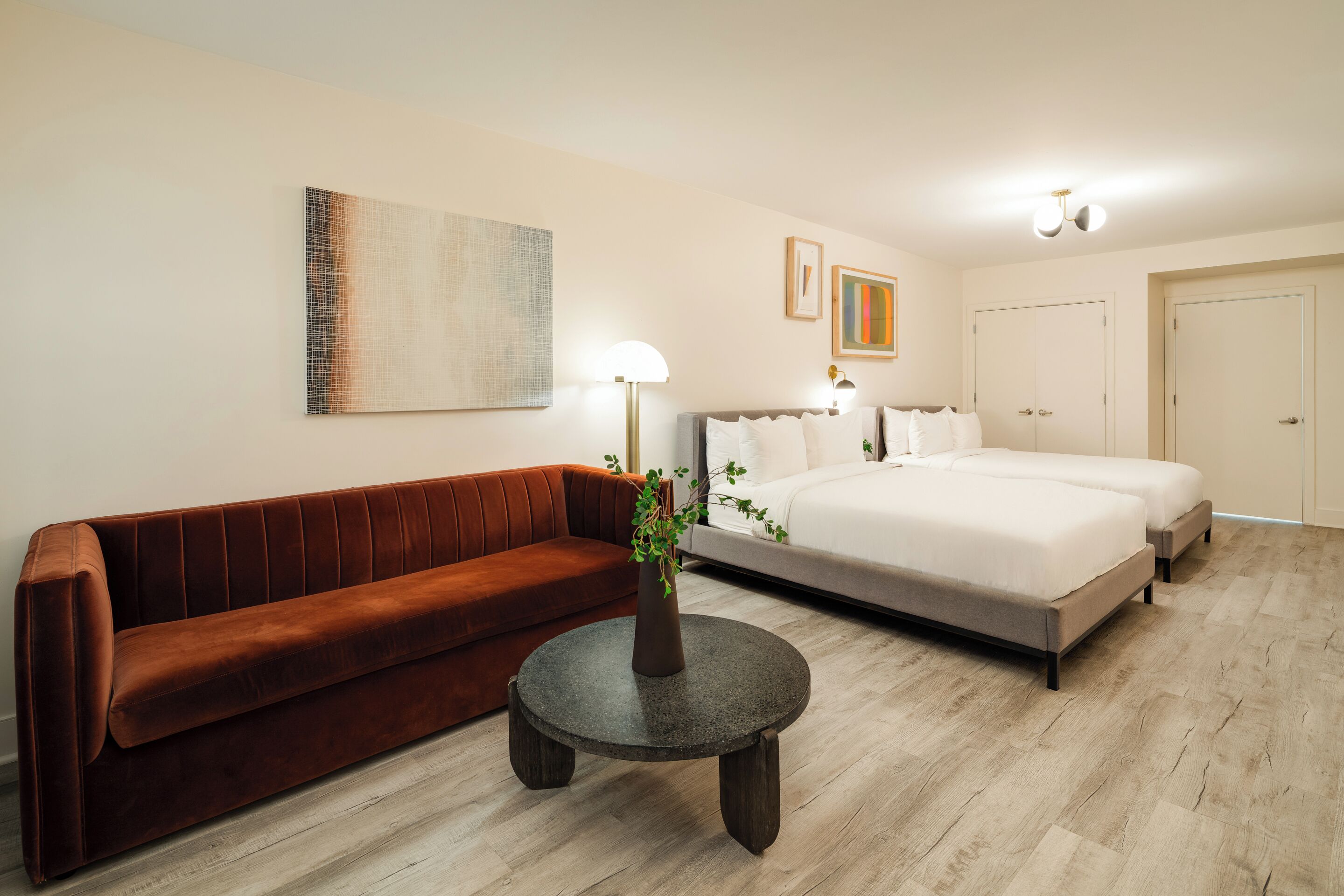 A modern hotel room with a brown sofa, a round table with a vase, a large bed, and a painting on the wall.