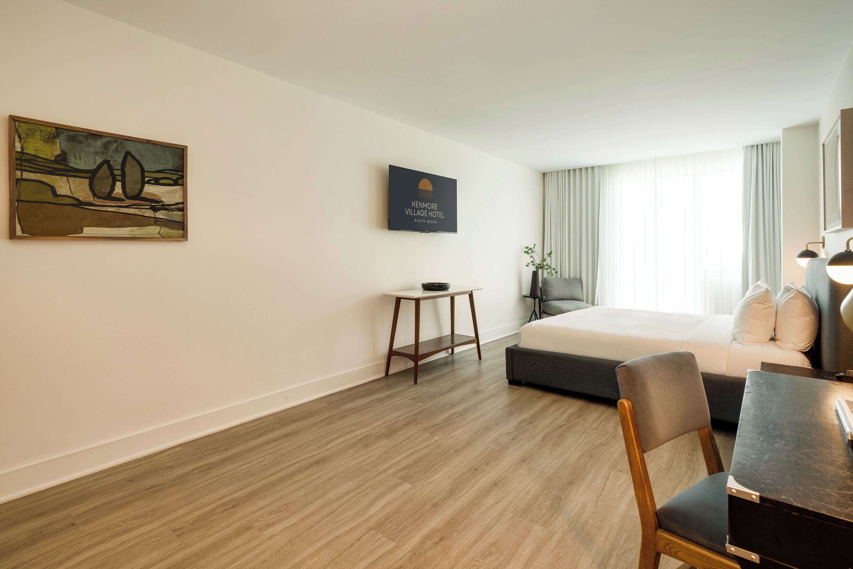 A hotel room with a bed, a desk, a chair, a small table, a painting, and a sign on the wall.