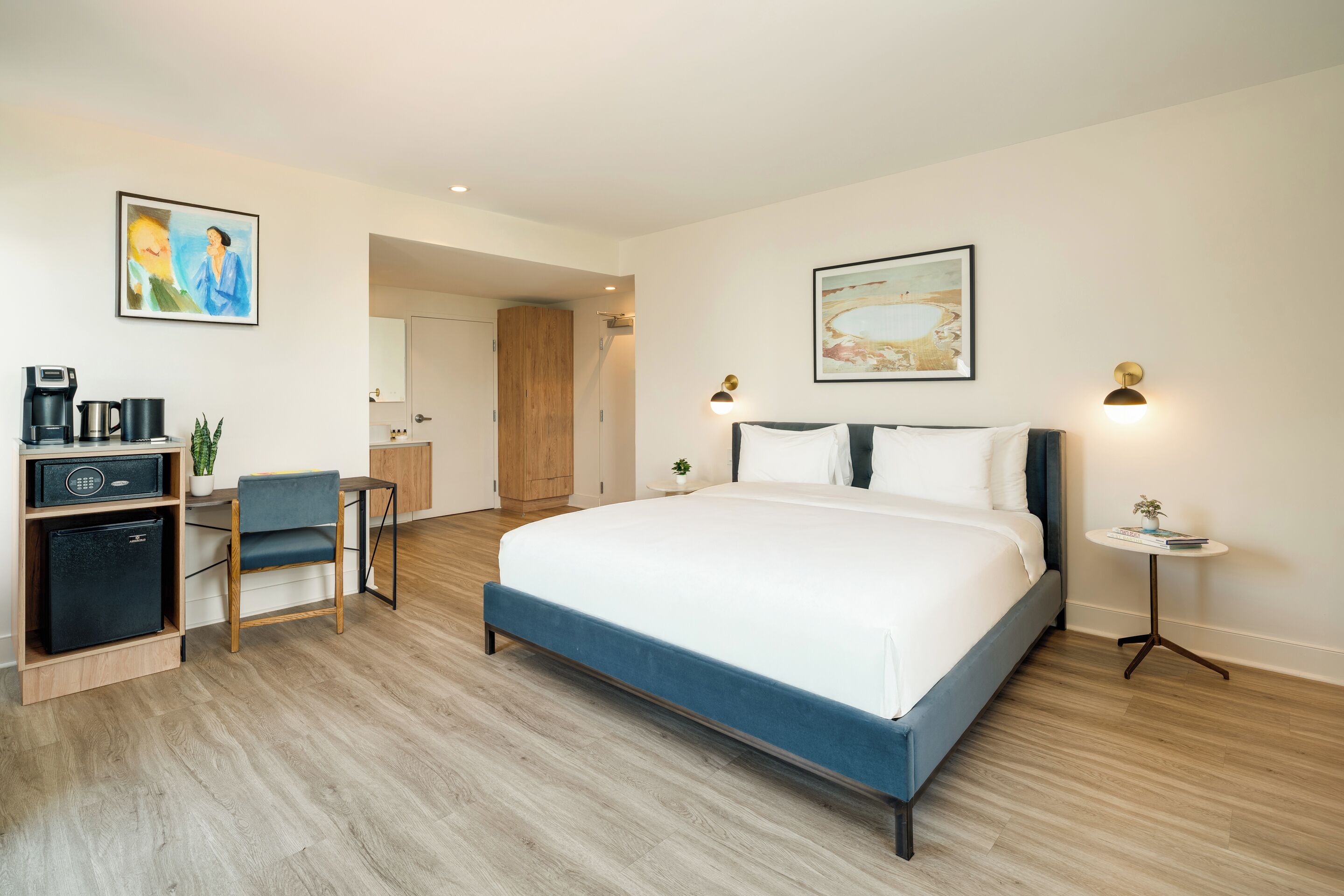 A modern hotel room with a large bed, a desk with a computer, a chair, and a small refrigerator.