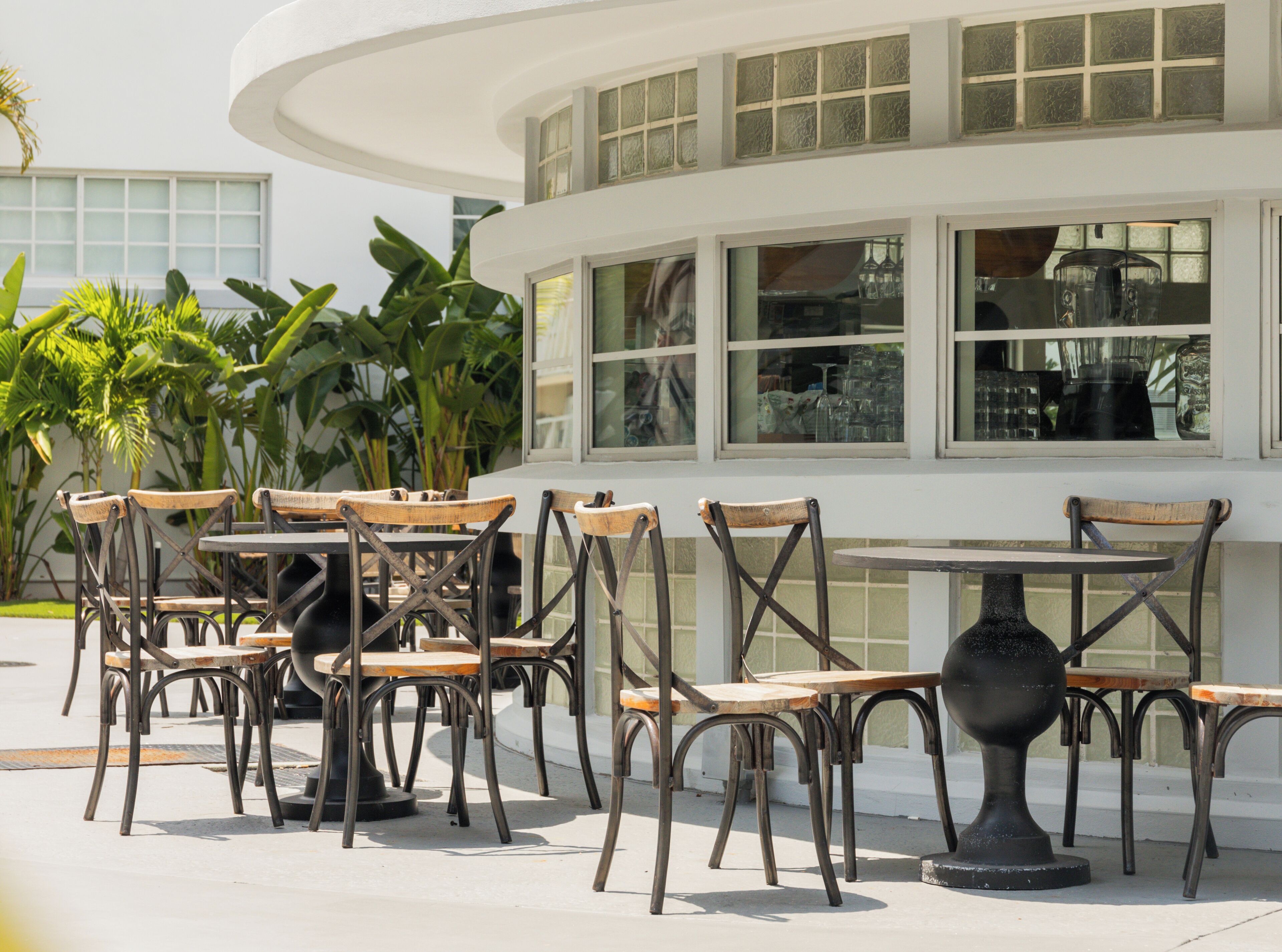 Een terras met tafels en stoelen, omgeven door grote ramen en groen.