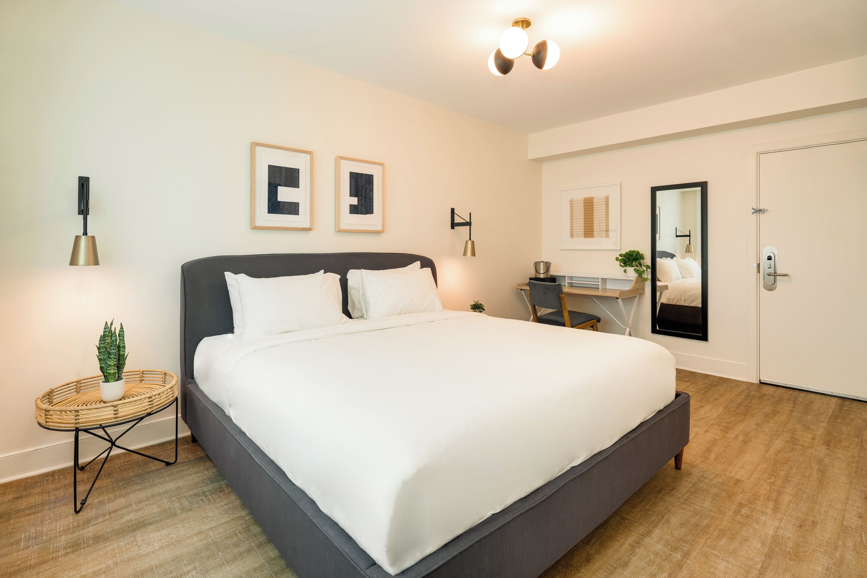 A modern bedroom with a large bed, a small side table with a plant, a desk with a chair, and a mirror on the wall.