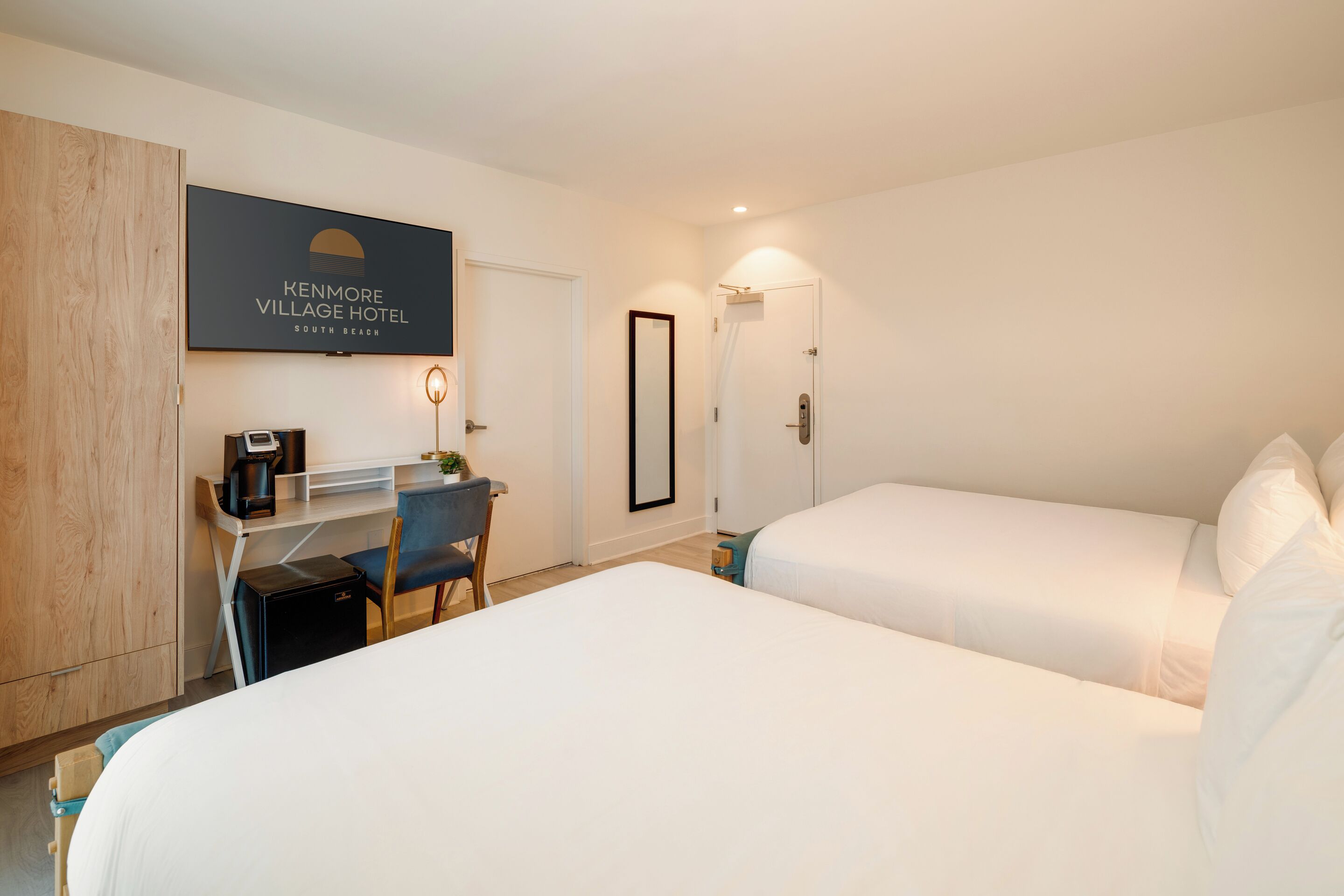 Ett hotellrum med två sängar, ett skrivbord med en dator, en kaffebryggare och en väggmonterad TV som visar hotellets namn.