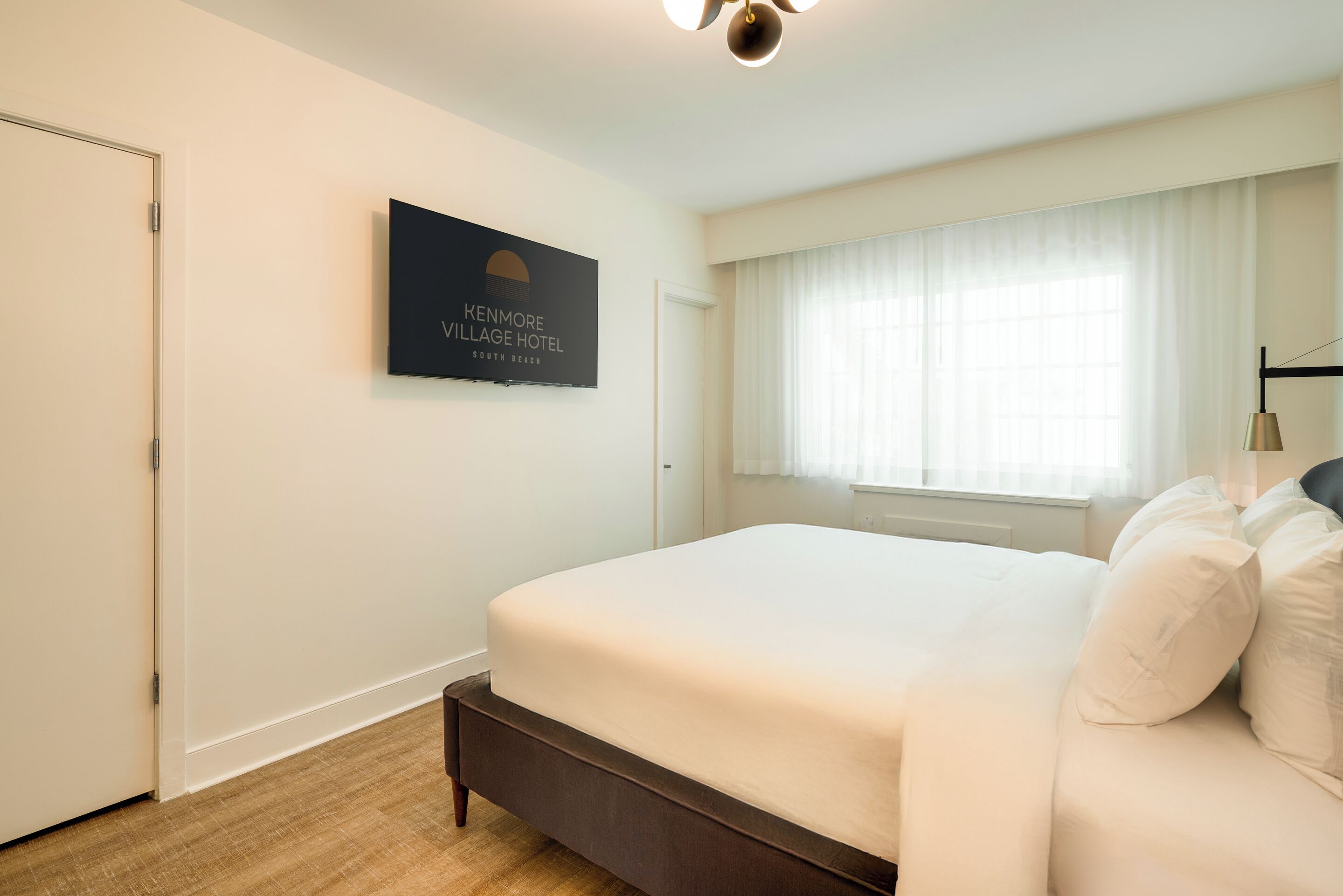 A hotel room with a large bed, a flat-screen TV mounted on the wall, a window with sheer curtains, and a wooden floor.