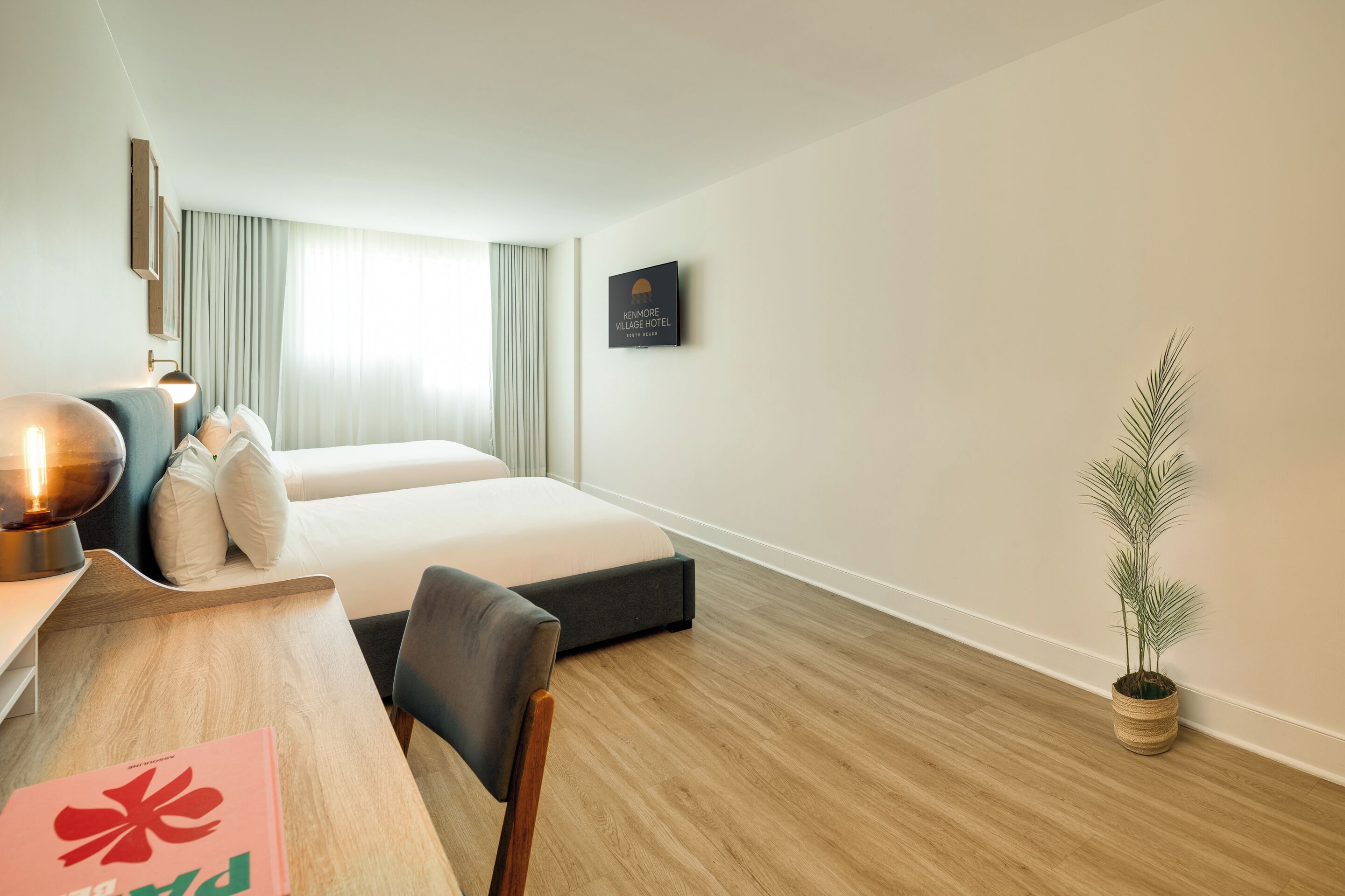 A hotel room with a bed, a desk, a chair, a lamp, a television, and a potted plant.