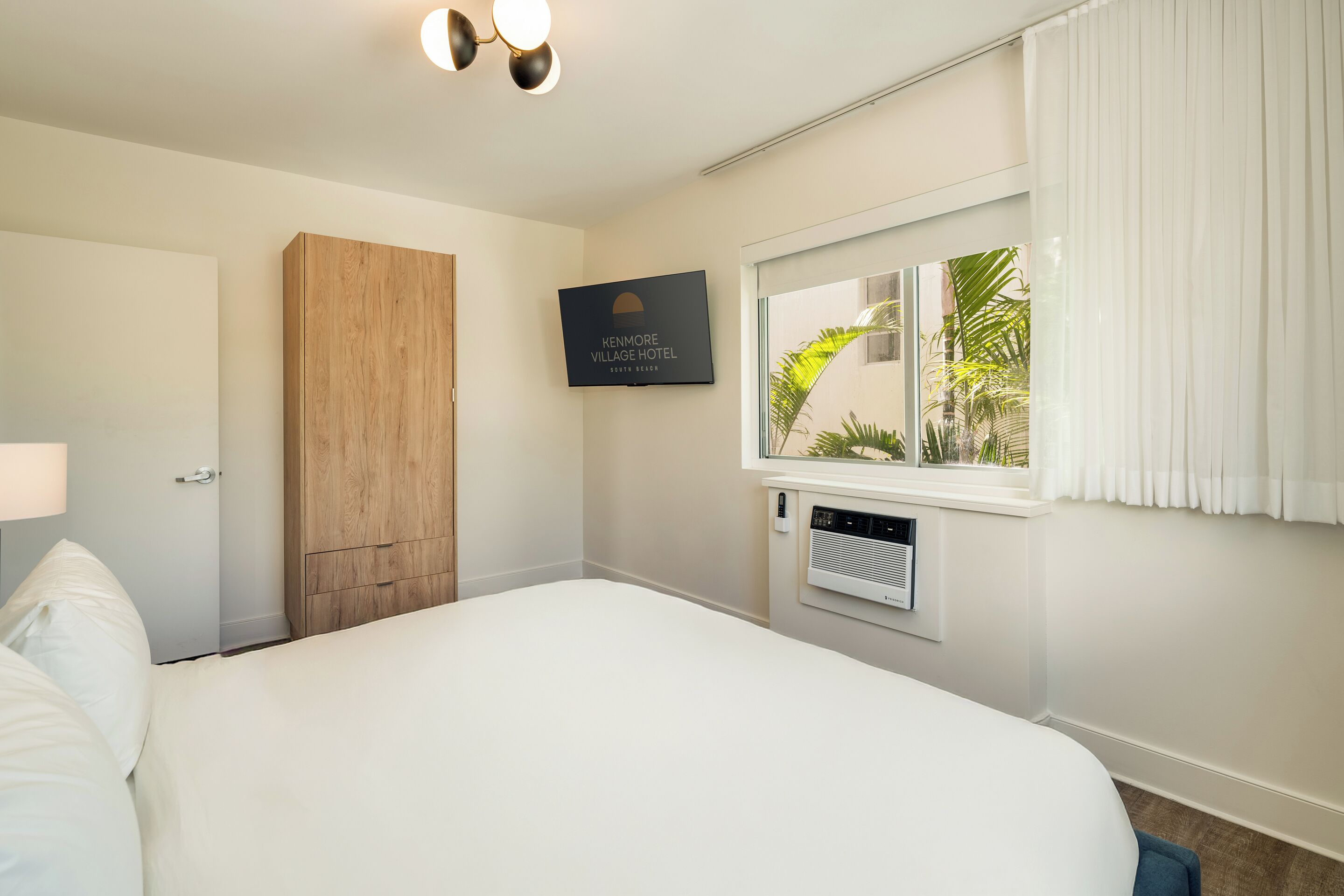 A hotel room with a large bed, a wooden wardrobe, a window with curtains, and a thermostat.